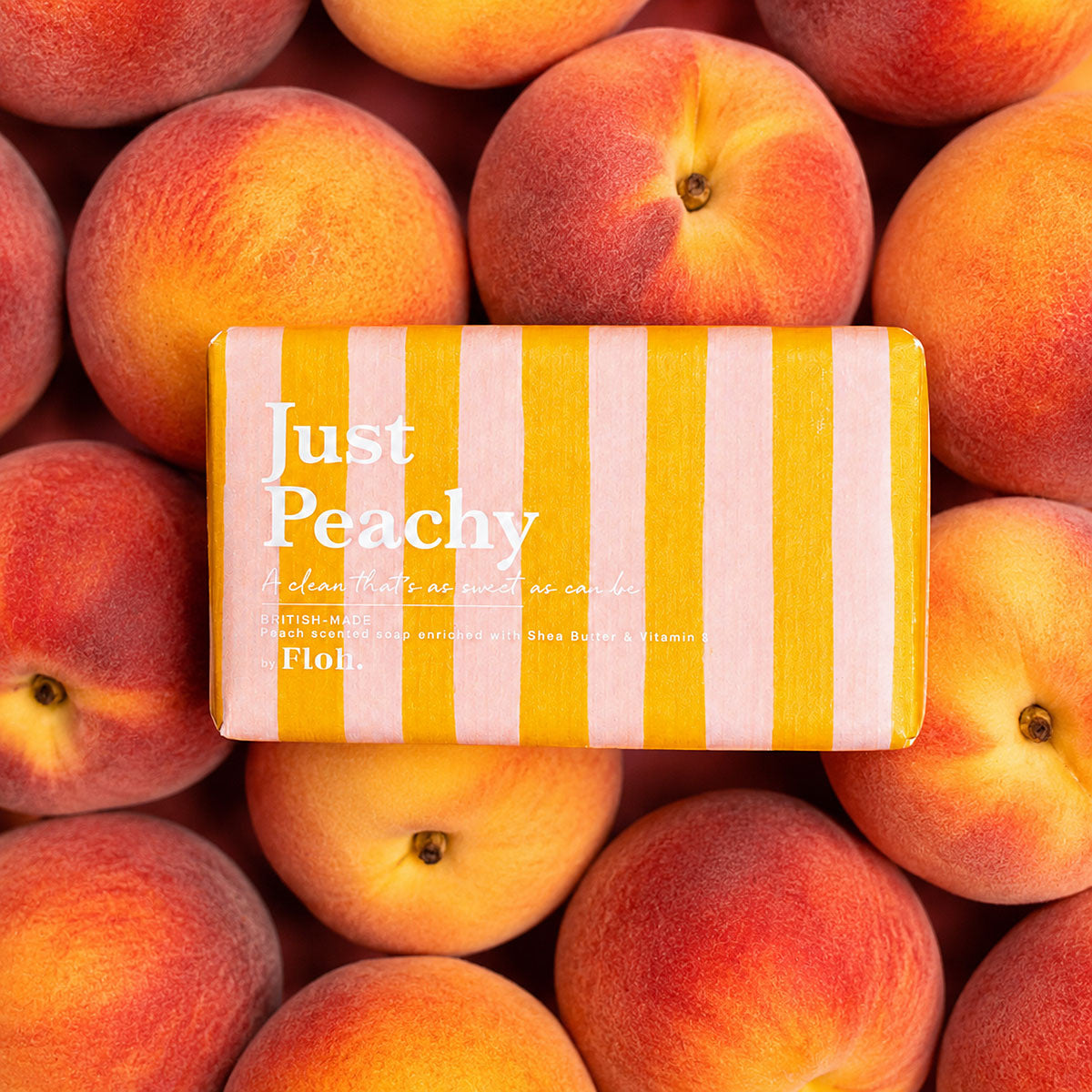 A rectangular bar of soap wrapped in yellow and white vertical stripes, placed on top of a dense arrangement of fresh peaches. The soap has the words 'Just Peachy' and the brand 'Flour' printed on it.