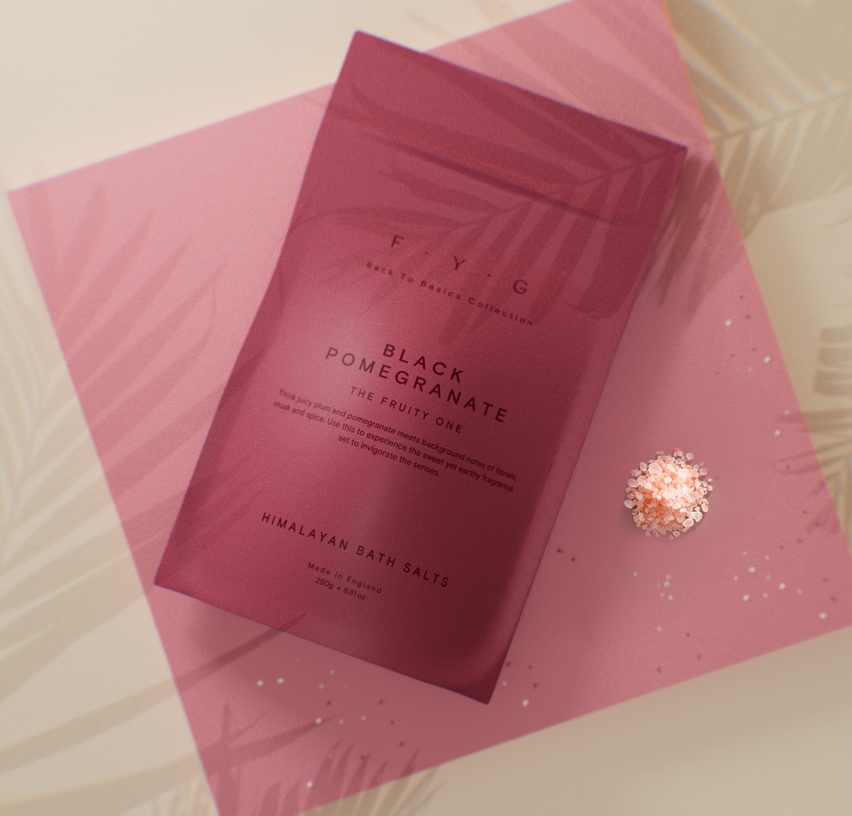 A pink foil packet of FYI G Himalayan Bath Salts, Black Pomegranate, placed on a pink leaf-patterned sheet with a small decorative rose-gold bead on a light beige background.