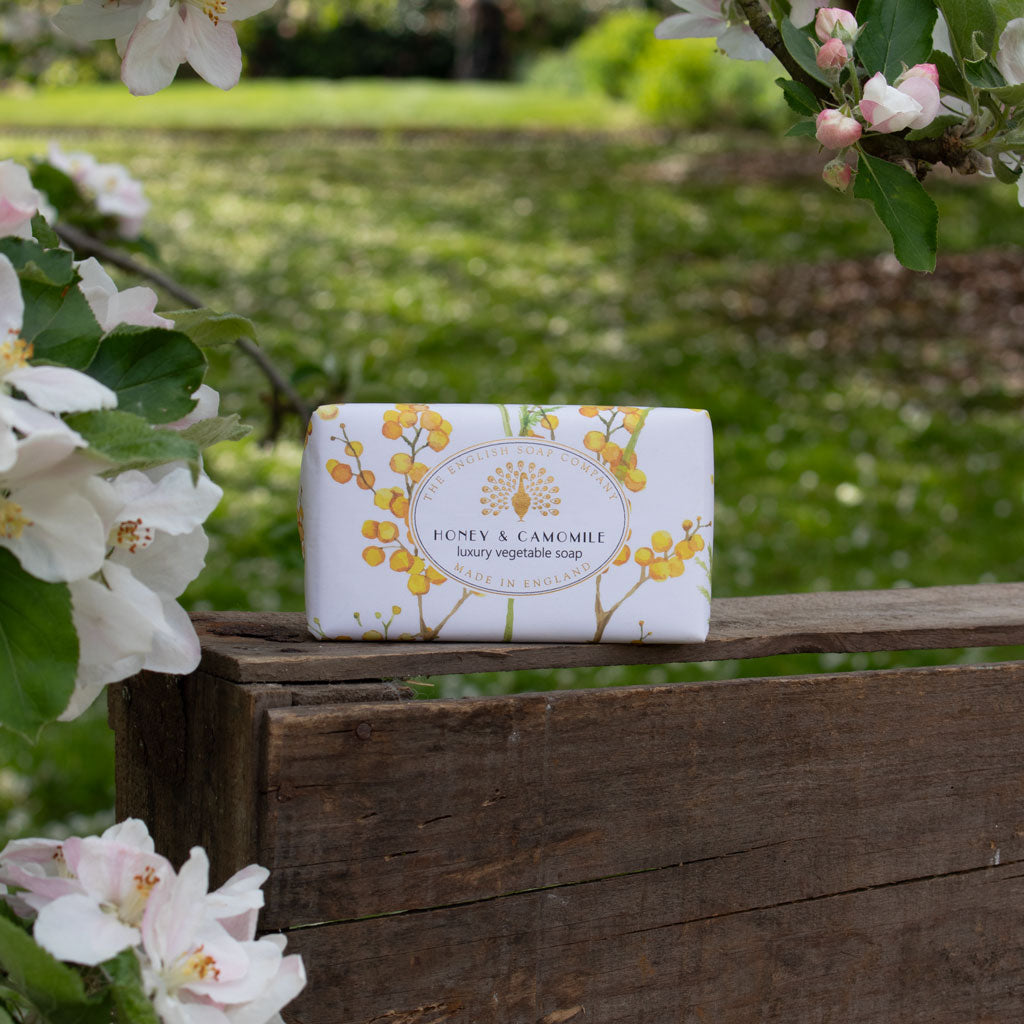 A rectangular bar of Honey & Camomile luxury vegetable soap sits on a rustic wooden fence outdoors, surrounded by blooming white flowers and a soft green garden background. - The Fig House