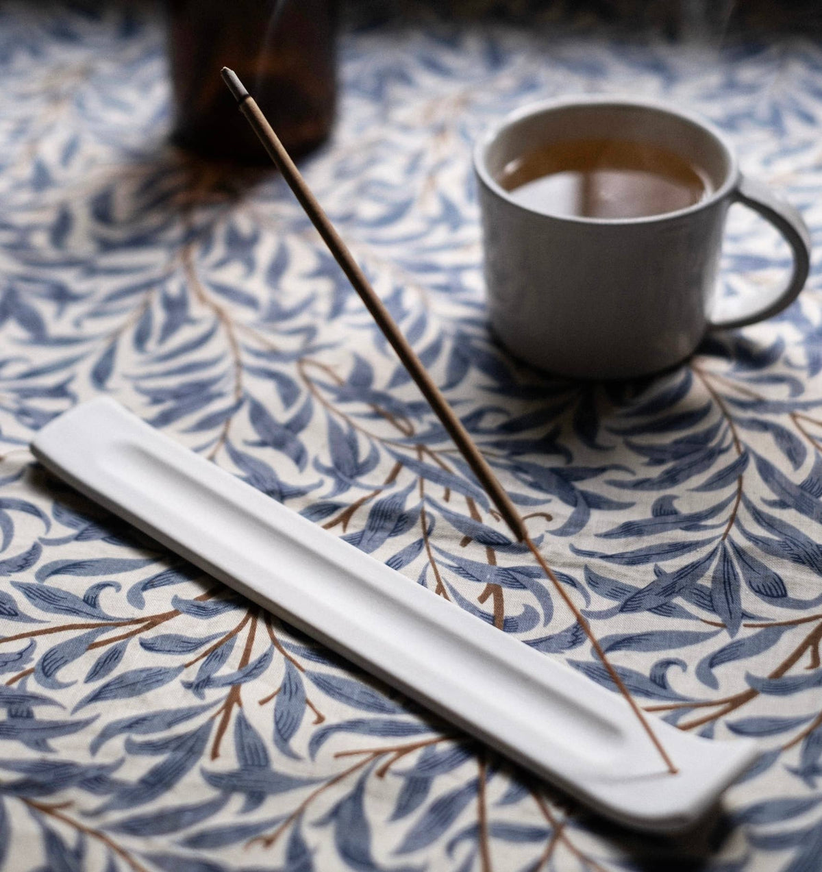 Incense stick on a white tray with a cup of tea on a patterned tablecloth
