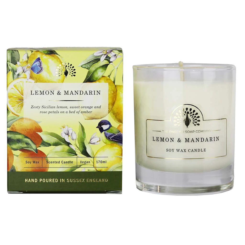 A lemon and mandarin scented soy wax candle by The Art of Living Company, shown next to its illustrated packaging box. The candle is in a clear glass jar with white wax, and the box features colorful illustrations of lemons, oranges, flowers, and birds. - The Fig House