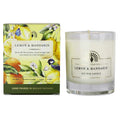 A lemon and mandarin scented soy wax candle by The Art of Living Company, shown next to its illustrated packaging box. The candle is in a clear glass jar with white wax, and the box features colorful illustrations of lemons, oranges, flowers, and birds. - The Fig House