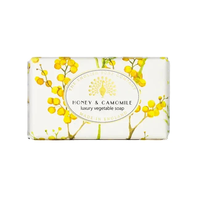 A rectangular bar of soap wrapped in white paper featuring a yellow floral pattern and a central white oval label that reads 'HONEY & CAMOMILE luxury vegetable soap MADE IN ENGLAND' by The English Soap Company.