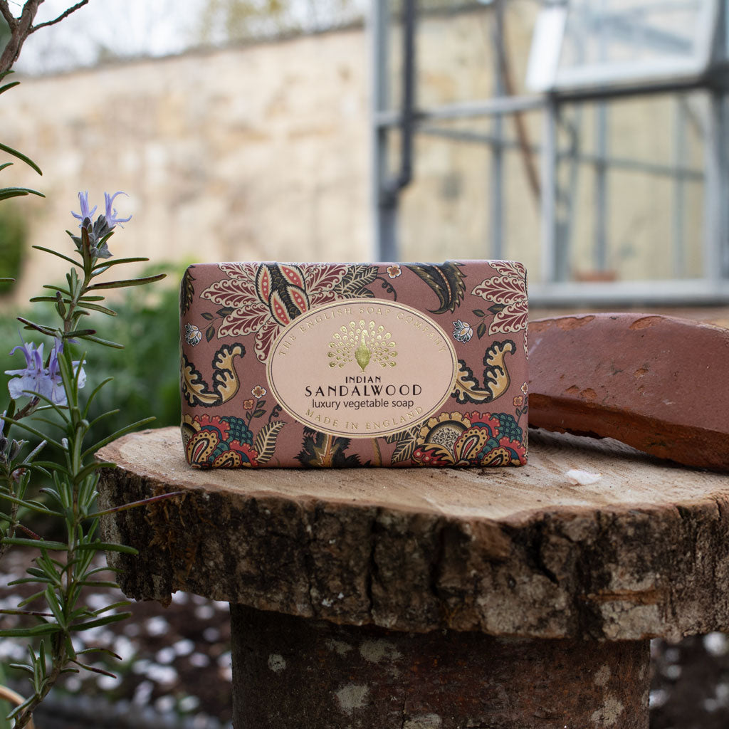 A rectangular bar of Indian Sandalwood luxury vegetable soap in ornate floral-patterned packaging sits on a rustic tree stump outdoors, with a matching soap bar resting behind it and garden plants blurred in the background. - The Fig House