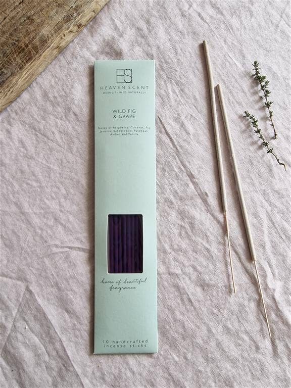 Heaven Scent Wild Fig & Grape incense sticks in their mint green packaging, displayed on a textured light pink linen fabric alongside two incense sticks and a small sprig of green herbs.
