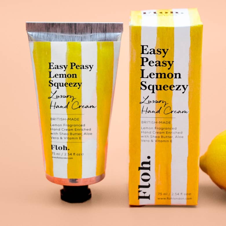 A squeeze tube and matching box of Floh. Easy Peasy Lemon Squeezy Luxury Hand Cream, featuring a yellow and white striped design with a black cap, set against a solid peach background with a lemon partially visible on the right.