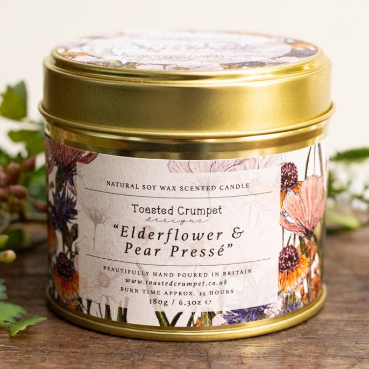 A round gold metal candle tin with a floral label reading 'Toasted Crumpet designs Elderflower & Pear Pressé', photographed on a wooden surface with soft greenery in the background.