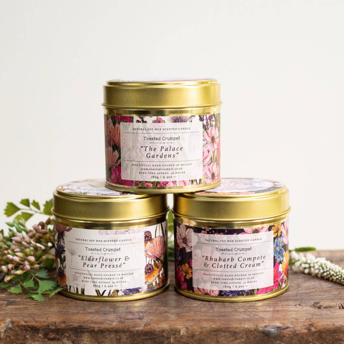 Three scented candles in decorative tins on a wooden surface with flowers in the background.