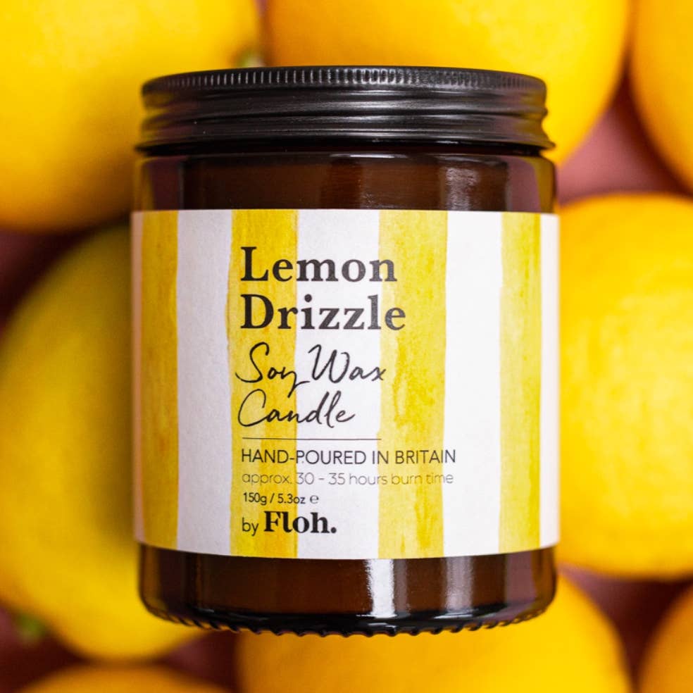 A jar of 'Lemon Drizzle' soy wax candle by Floh. The amber glass jar has a black lid and a white label with yellow stripes, set against a background of fresh yellow lemons.