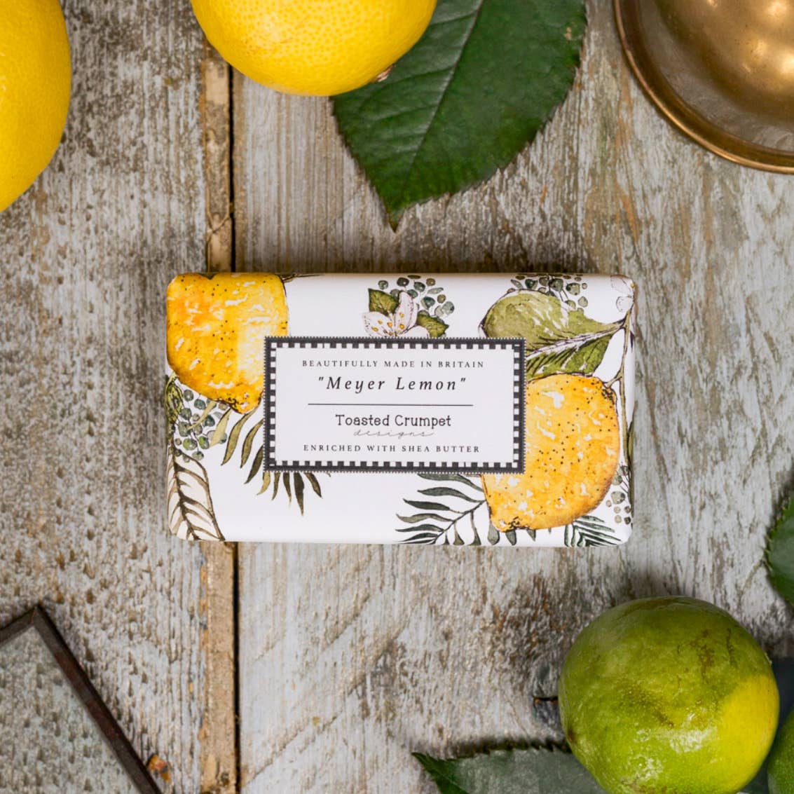 Bar of soap with 'Meyer Lemon' packaging on a wooden surface with lemons and limes.
