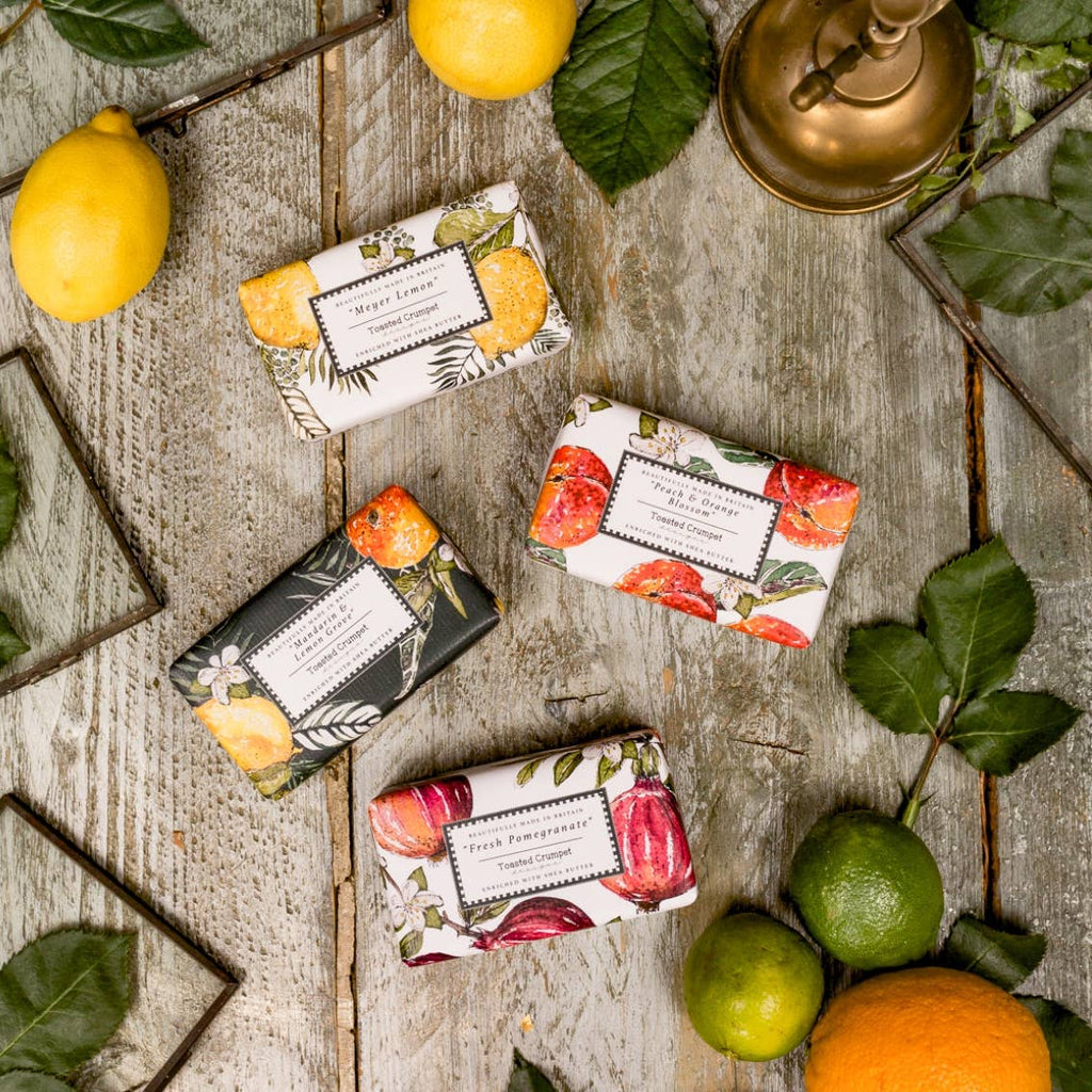 Four soap bars with fruit-themed designs on a wooden surface with lemons and limes.
