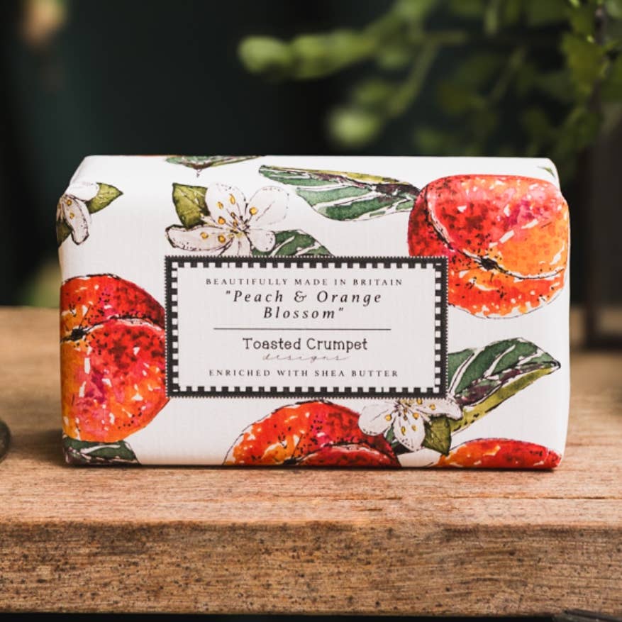 A rectangular bar of soap wrapped in white paper printed with peach and orange blossom illustrations, resting on a rustic wooden surface with a blurred green outdoor background.