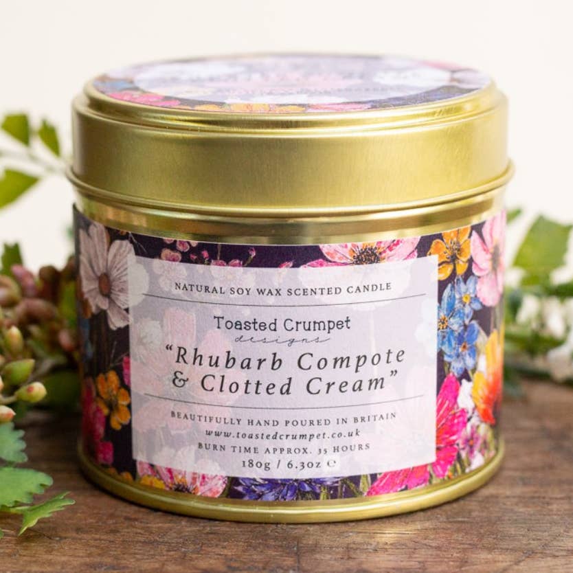 A round metal tin candle with a gold lid and a colorful floral label reading 'Toasted Crumpet designs Rhubarb Compote & Clotted Cream'. The tin sits on a wooden surface with green leaves around it.