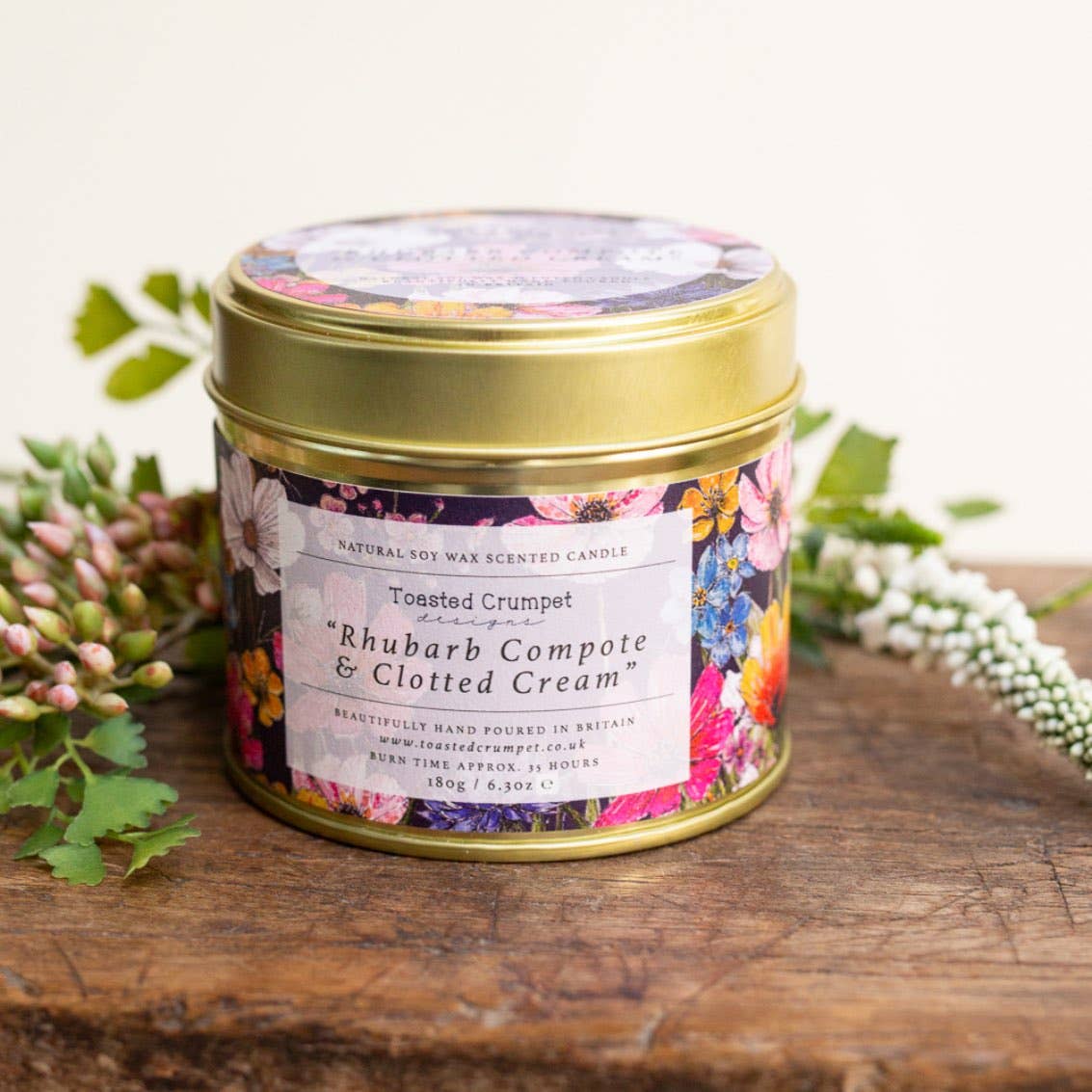 A round gold metal candle tin with a colorful floral label and lid, placed on a rustic wooden surface with flowers around it. The label reads 'Natural Soy Wax Scented Candle' and 'Rhubarb Compote & Clotted Cream'.