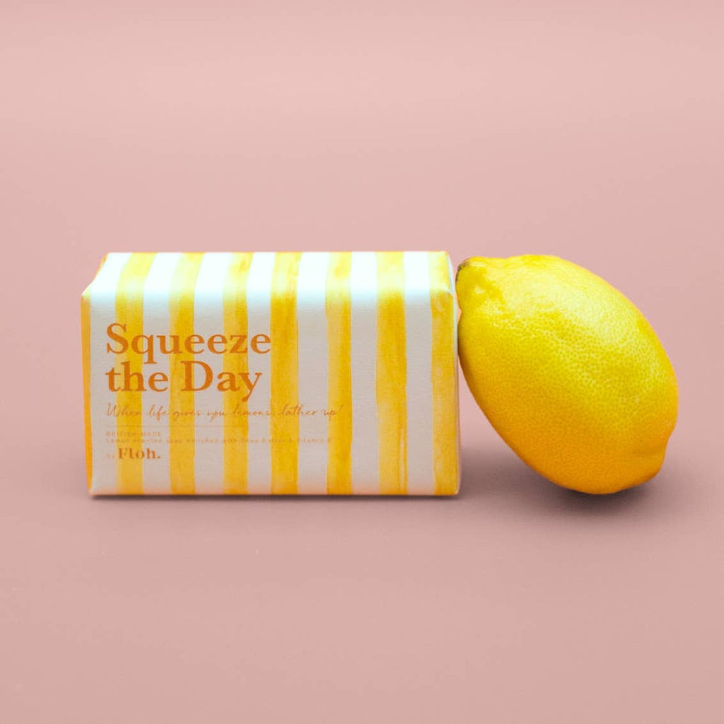 Bar of soap with yellow and white striped packaging labeled 'Squeeze the Day' next to a lemon on a pink background