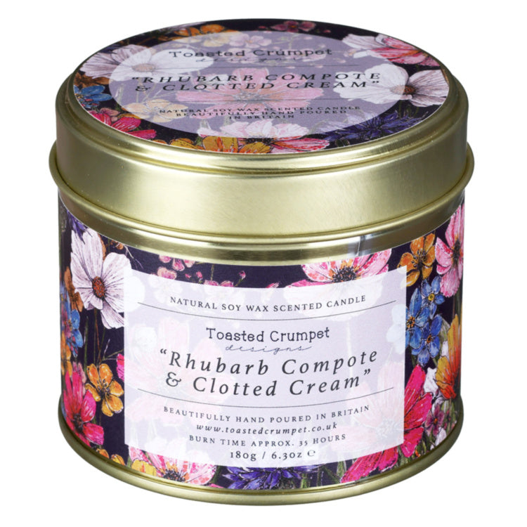 A cylindrical tin candle with a gold metal lid and a colorful floral label, shown on a plain white background. The label reads 'Toasted Crumpet' and 'Rhubarb Compote & Clotted Cream'.