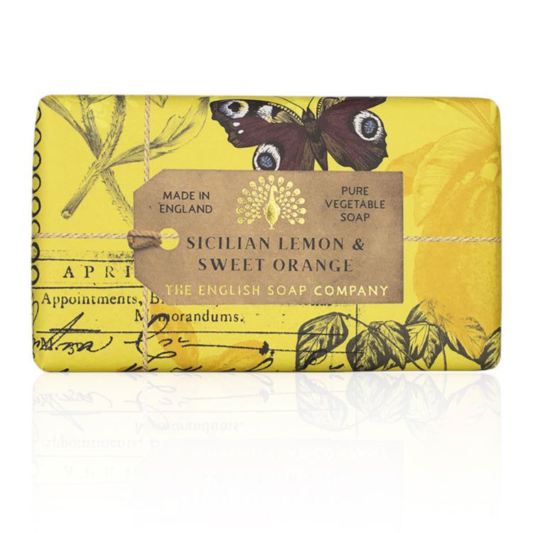 A rectangular bar of soap wrapped in yellow paper with botanical illustrations, a butterfly, and script, featuring a brown paper label that reads 'SICILIAN LEMON & SWEET ORANGE' and 'THE ENGLISH SOAP COMPANY'.